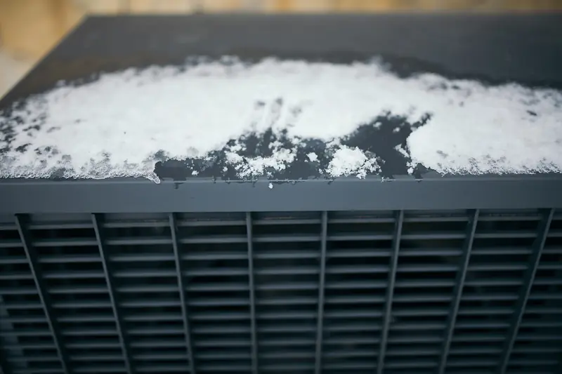 Snow and slush lying on the flat dark grey top of an outdoor unit, above its slatted coil guard